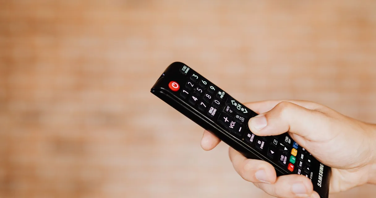 Mão segurando um controle remoto de televisão diante de uma parede de tijolos desfocada, ilustrando o cache da caixa de streaming