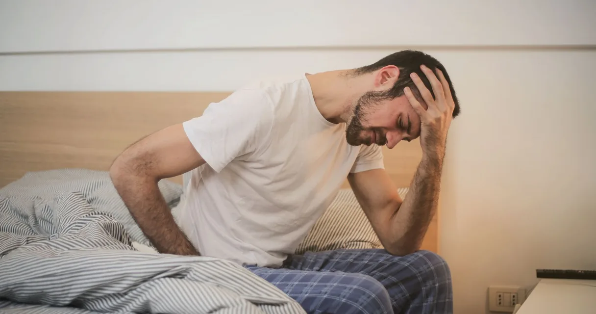 Homem sentado na beira da cama pela manhã, curvado e com a mão na cabeça